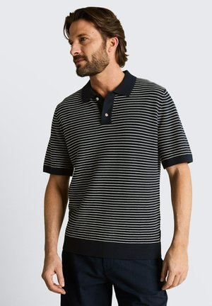 Man met baard die een zwart-wit horizontaal gestreept poloshirt met korte mouwen en donkere broek draagt, staand tegen een effen lichte achtergrond.