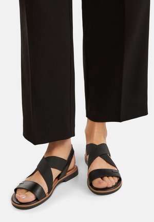 Nohy v čiernych kožených a textilných sandáloch s križujúcimi sa popruhmi sú spárované s čiernymi širokými nohavicami na bielom pozadí.