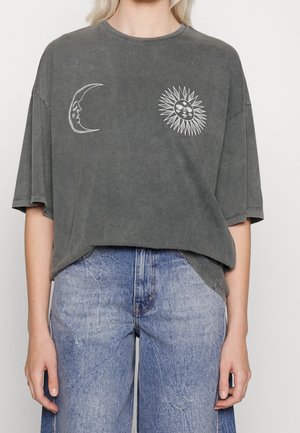 Kobieta nosząca luźny ciemnoszary t-shirt z białymi grafikami twarzy słońca i księżyca, w połączeniu z jasnoniebieskimi jeansami z wysokim stanem.