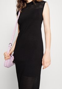 Robe noire sans manches avec un col montant; présente une texture côtelée sur la partie supérieure et une section inférieure unie. Accentuée par un sac à main rose.