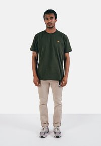 T-shirt en coton vert olive avec un petit logo d'éléphant jaune sur la poitrine, associé à un pantalon beige et des baskets multicolores.