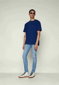 Jovem de camiseta oversized azul, jeans skinny claros rasgados, ténis brancos de cano alto e óculos de sol com lente azul, posando contra um fundo simples.