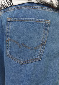 Zadní kapsa na džínách světle modré barvy s kontrastním žlutým prošitím. Kapsa má design zahnuté linie. Textura je mírně strukturovaná.