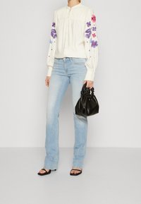 Vit blus med blommönster i lila och rosa, kombinerad med ljusblå utsvängda jeans och en liten svart texturerad handväska.