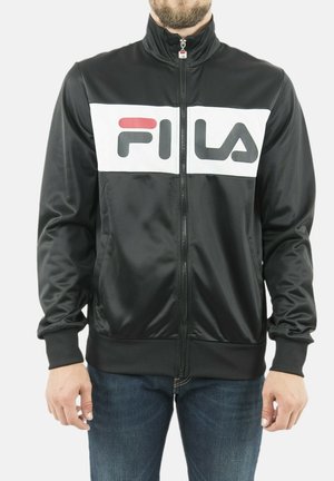 Uomo che indossa una giacca nera con zip con un pannello bianco sul petto che mostra il logo FILA in nero e rosso.