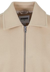 Beige fleecejakke med spiss krage, frontlynlås og en myk tekstur. Merket "URBAN CLASSICS," størrelse XS.