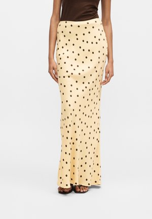 Μπεζ μακριά φούστα με μαύρα πουά, συνδυασμένη με καφέ αμάνικη μπλούζα και μαύρα σανδάλια, που στέκεται σε απλό λευκό φόντο.