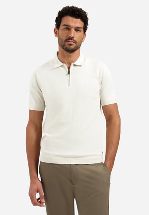 Mann mit kurzen lockigen Haaren und Bart, der ein weißes kurzärmeliges Polo mit Reißverschluss und olivgrüne Hose trägt, steht mit einer Hand in der Tasche.