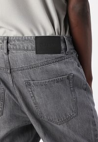 Grå denim jeans med en teksturert finish, med en svart skinn merkepute og to baklommer med sømdetaljer.