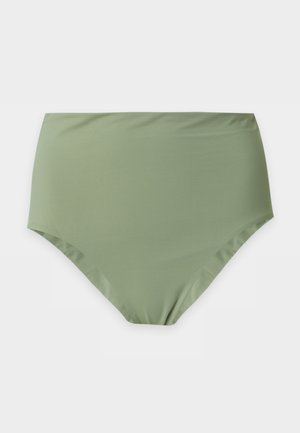 Femme debout portant un maillot de bain deux pièces vert olive avec un bas taille haute et des tongs noires sur un fond blanc uni.