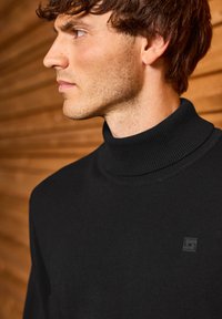 Homme aux cheveux bruns courts portant un pull noir à col roulé, se tenant contre un mur en lattes de bois, regardant vers la gauche.