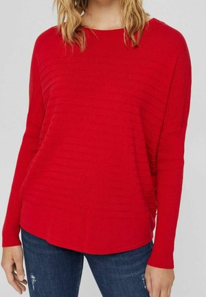 Femme portant un pull rouge vif à manches longues avec des détails en côtes horizontales, associé à un jean bleu foncé déchiré, se tenant devant un fond uni.