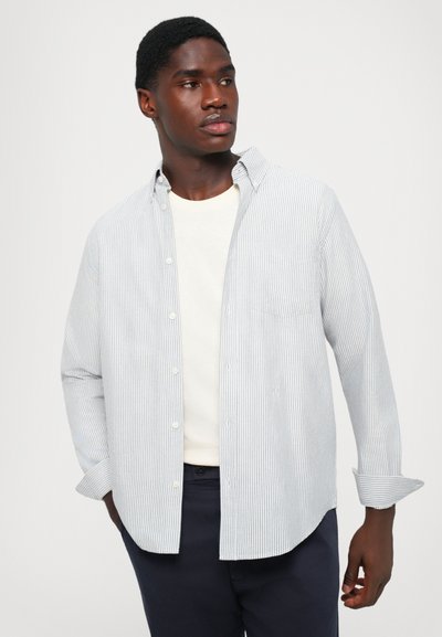 Light blue and white striped button-up shirt with a collar, long sleeves, and a chest pocket, worn over a cream-colored shirt.