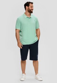 Jasnozielona koszulka polo z krótkim rękawem i kołnierzykiem, zestawiona z ciemnoniebieskimi szortami. Model nosi białe trampki.