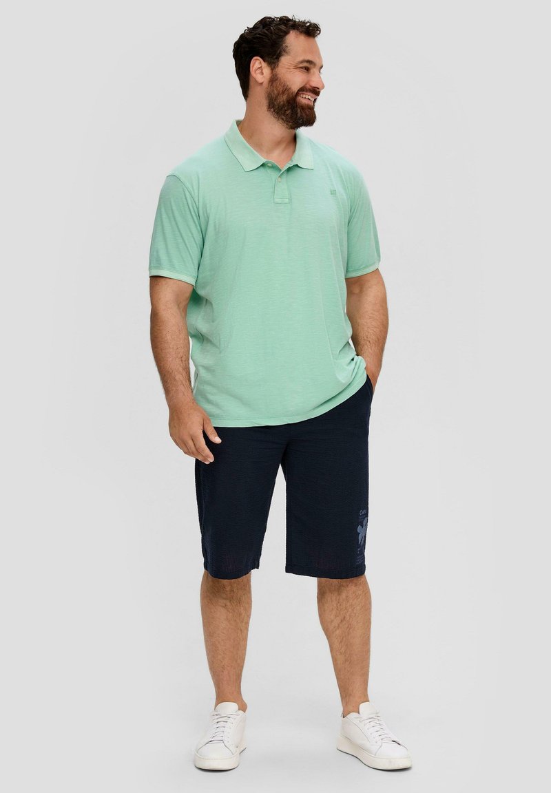 Jasnozielona koszulka polo z krótkim rękawem i kołnierzykiem, zestawiona z ciemnoniebieskimi szortami. Model nosi białe trampki.