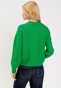 Groene gebreide trui met een ronde hals, doorgezakte schouders en geribbelde zoom. Zachte textuur en losse pasvorm, gecombineerd met donkere spijkerbroek.