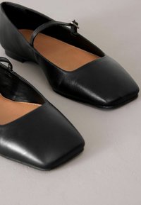 Zapatos planos de cuero negro con punta cuadrada, una delgada tira sobre el empeine y una textura suave. La suela es plana y resistente.
