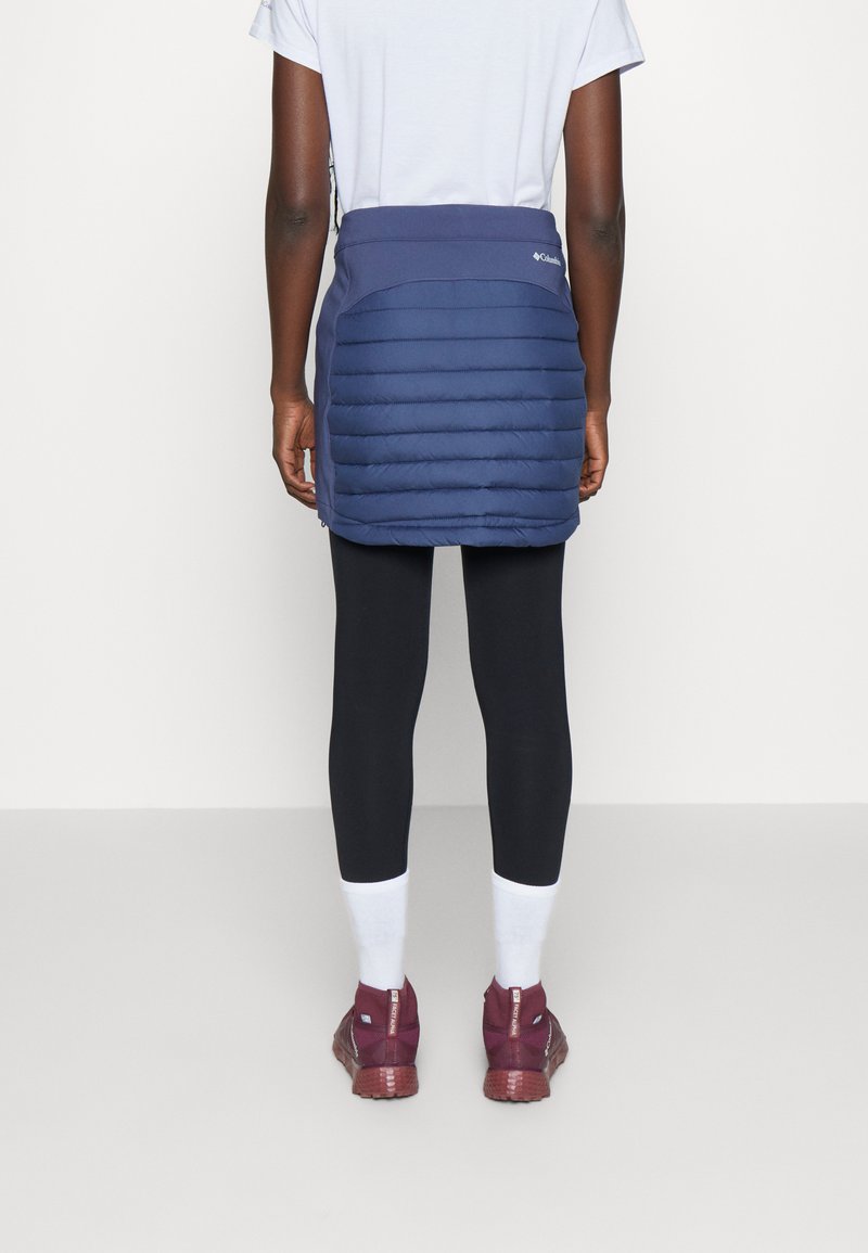 Granatowa spódnica puchowa z poziomym pikowaniem, zestawiona z czarnymi legginsami i bordowymi sneakersami. Zaprojektowana z myślą o ciepłości i komforcie.