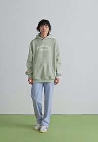En ung person står i en ljusgrön hoodie med "New Balance Athletics", ljusblå byxor och vita sneakers framför en enfärgad vit och grön bakgrund.