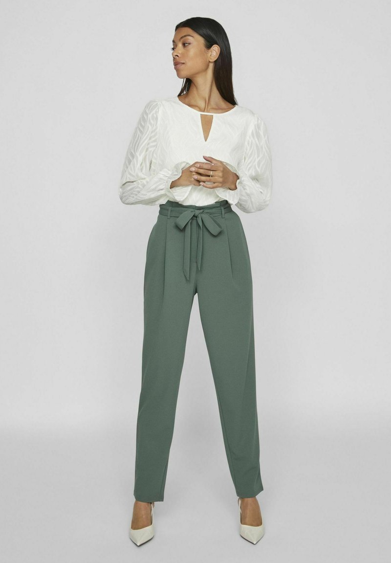 Witte blouse met gestructureerde mouwen en een sleutelgatdetail, gedragen met high-waisted, groene broek en crèmekleurige puntige hakken.