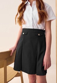 Jeune personne aux longs cheveux roux portant une chemise blanche à manches courtes boutonnée et rentrée dans une jupe noire plissée, debout à côté d'une chaise en bois.