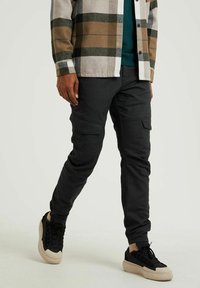 Man die loopt in zwarte cargobroek, zwarte sneakers met dikke beige zolen, een donkergroen shirt, en een bruin-groen geruit jack.