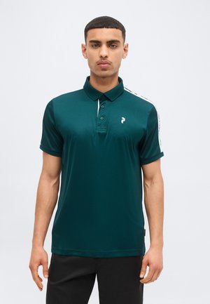 Jeune homme aux cheveux courts portant un polo vert foncé avec une bande blanche sur l'épaule et un petit logo, debout devant un fond clair uni.