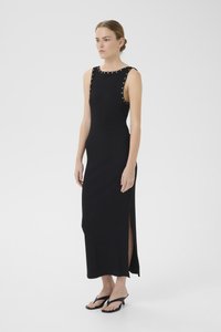 Vestido maxi preto sem mangas com fenda lateral, apresentando rebites metálicos ao longo do decote. Textura suave com uma silhueta ajustada. Chinelos pretos.