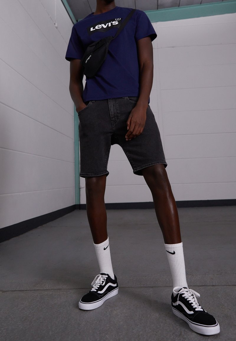 T-shirt Levi's bleu marine, short en denim noir, chaussettes blanches Nike et baskets Vans noires. Tenue décontractée avec un sac bandoulière noir compact.