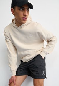 Beige hooded sweatshirt with white Jordan logo, paired with black shorts featuring a white logo, worn with a black cap; indoor setting.