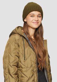 Olijfgroene pufferjas met een capuchon, voorzien van een diamantgequilt patroon en een grijze voering. Gecombineerd met een donkergroene ribgebreide beanie.