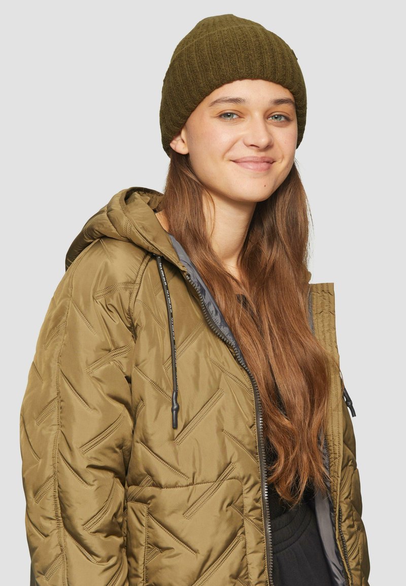 Olijfgroene pufferjas met een capuchon, voorzien van een diamantgequilt patroon en een grijze voering. Gecombineerd met een donkergroene ribgebreide beanie.