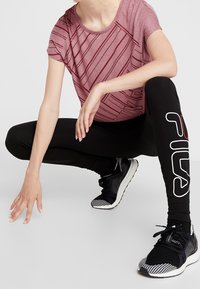 Czerwona koszulka sportowa w paski, czarne legginsy z białym logo oraz czarne buty do biegania z paskowanymi akcentami. Modelka przykucnęła na jasnym tle.