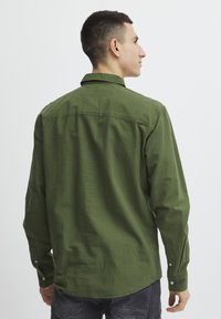 Camisa verde de botones, hecha de tela texturizada, con cuello, mangas largas y dobladillo curvo. La vista trasera muestra las costuras de los hombros y los botones.