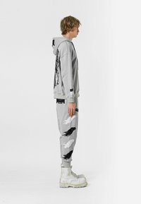 Joven con sudadera gris y pantalones con diseños de alas en blanco y negro, de pie de lado, llevando botas blancas de cordones sobre un fondo blanco.