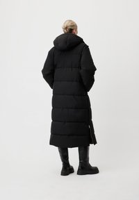 Personne aux cheveux blonds en chignon portant un long manteau en duvet noir avec capuche et des bottes noires jusqu'aux genoux, se tenant contre un fond blanc uni.