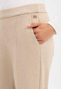 Beige Hose mit strukturiertem Muster, ausgestattet mit einer Fronttasche und einem metallischen Logo-Akzent am Bund für Details.