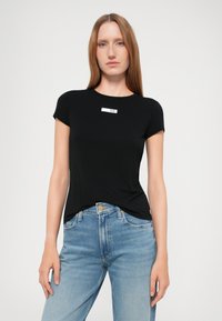 Zwarte T-shirt met korte mouwen en een ronde halslijn, voorzien van een wit label met "EA7" op de borst. Gepaard met lichtblauwe denim jeans.