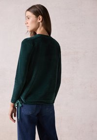 Haut en velours vert à manches longues avec un lien à la taille, associé à un jean bleu foncé. Texture douce et coupe décontractée.