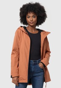 Giacca resistente all'acqua di colore ruggine con cappuccio, chiusura con zip e polsini elasticizzati. Abbinata a una maglia nera e jeans blu.