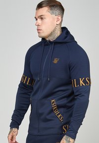 Felpa con zip di colore blu navy con dettagli e branding dorati sulle maniche. Realizzata in un tessuto liscio ed elastico, con tasca a marsupio e cappuccio.