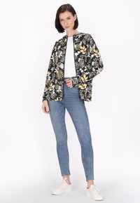 Jas met bloemenprint in zwart, met witte, gele en blauwe bloemen; gedragen over een witte top en gecombineerd met blauwe jeans en witte sneakers.