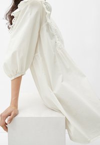 Weißes Baumwollkleid mit puffärmeln, versehen mit Lochstickerei und Rüschenakzenten entlang des Ausschnitts, das sanft über einem weißen Sockel hängt.