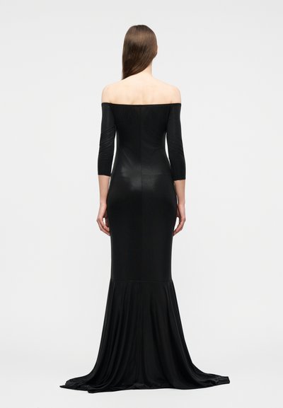 Γυναίκα με μακριά μαλλιά που φοράει στενή μαύρη τουαλέτα χωρίς τους ώμους, με μανίκια τριών τετάρτων και μακριά ουρά, στέκεται γυρισμένη προς τα πίσω σε απλό φόντο.