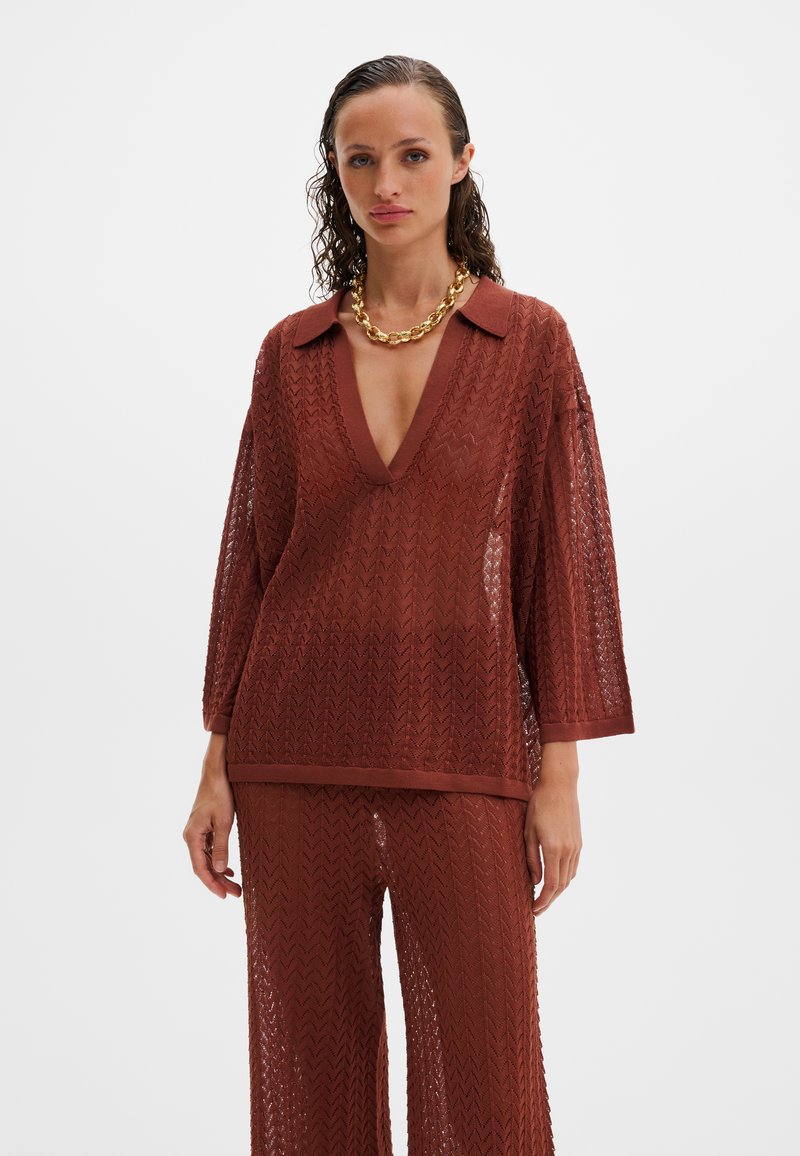 Γυναίκα που φοράει σετ από πλεκτή μπλούζα και παντελόνι σε χρώμα σκουριάς με ζιγκ ζαγκ σχέδιο, βαθύ ντεκολτέ σε σχήμα V και χρυσό χοντρό αλυσίδα λαιμού, σε λευκό φόντο.