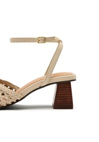 Sandalias trenzadas beige con una correa de tobillo ajustable y un tacón de bloque geométrico marrón. Textura suave con diseño minimalista.