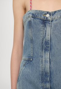 Denim kombinezon v bledi modri barvi s strukturiranim dizajnom. Ima sprednje zapiranje na gumb, dva stranska žepa in teksturirano tkanino.