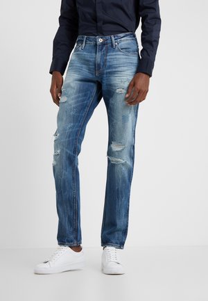 Homme portant un jean bleu usé, des baskets blanches et une chemise à manches longues foncée, debout devant un fond uni clair.