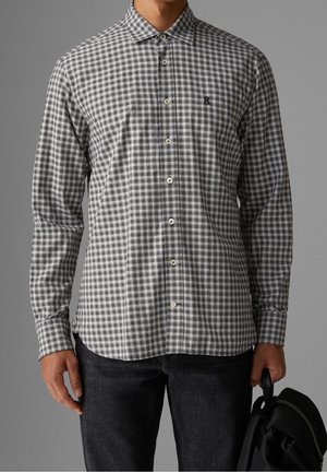 Homme portant une chemise à carreaux grise et blanche boutonnée avec un pantalon noir, tenant un sac à dos noir par la poignée.