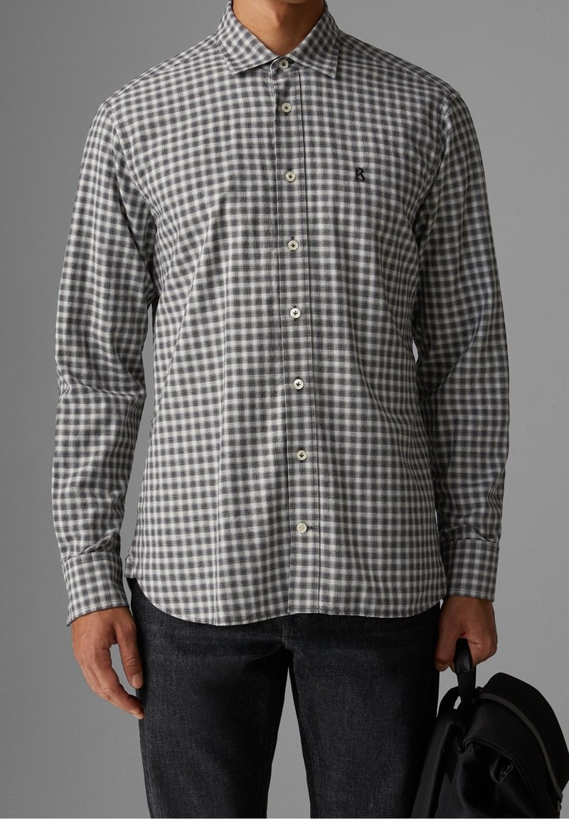 Homme portant une chemise à carreaux grise et blanche boutonnée avec un pantalon noir, tenant un sac à dos noir par la poignée.